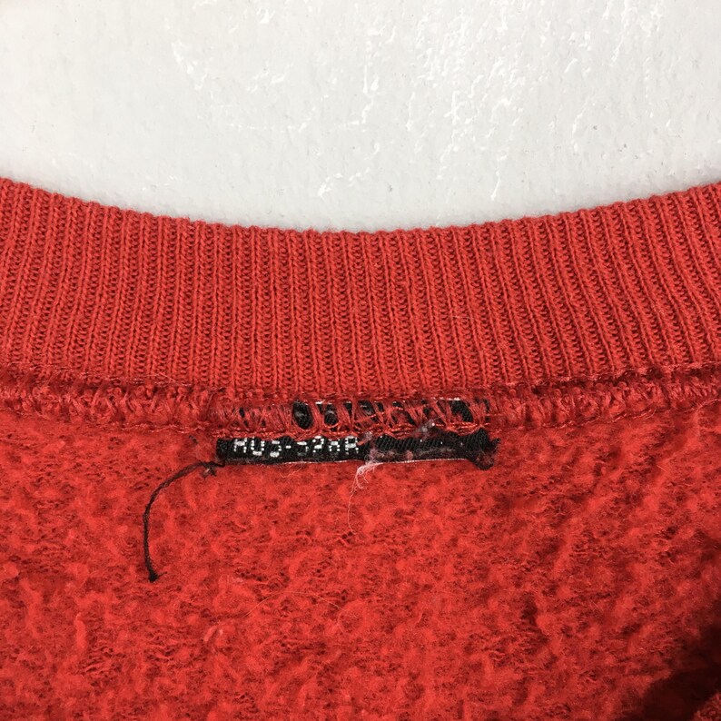 Peut inclure: Gros plan sur un col de sweat-shirt rouge. Le col est c&ocirc;tel&eacute; et le tissu a un aspect textur&eacute;. Une petite &eacute;tiquette noire avec du texte blanc est visible pr&egrave;s du col.