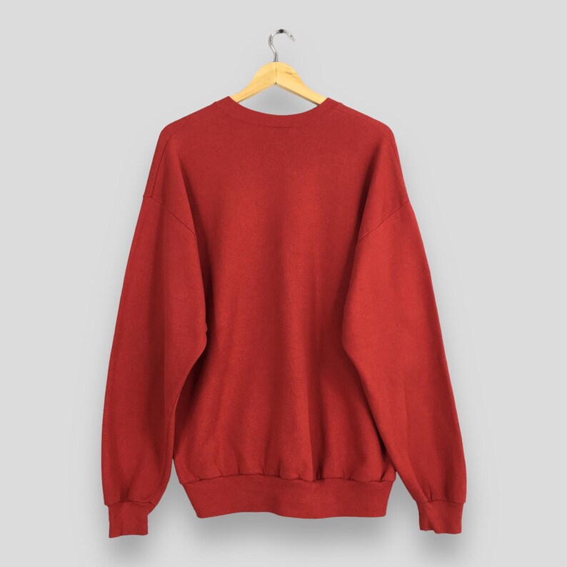 Peut inclure: Un sweat-shirt rouge uni est suspendu &agrave; un cintre en bois sur un fond gris clair. Le sweat-shirt a un col rond, des manches longues et des poignets et un ourlet c&ocirc;tel&eacute;s. Le tissu semble &ecirc;tre un mat&eacute;riau doux et de poids moyen.