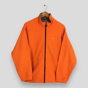 May include: Vibrant orange fleece jacket featuring a full-length black zip and a small white Nike logo on the chest. The jacket has a high collar and a grey lining. It is suspended from a wooden hanger.