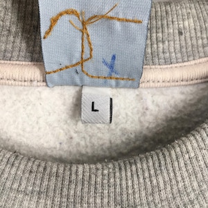 May include: Close-up of a gray sweatshirt with a light pink neckline. A light blue tag with gold stitching and a blue 'X' is visible. A white tag with a black 'L' is also present.