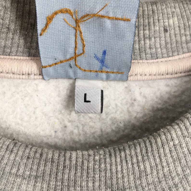 May include: Close-up of a gray sweatshirt with a light pink neckline. A light blue tag with gold stitching and a blue 'X' is visible. A white tag with a black 'L' is also present.