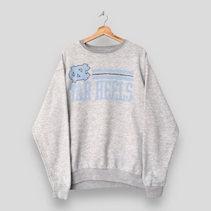 Op de afbeelding: Een grijze sweatshirt met een lichtblauw en zwart ontwerp. De sweatshirt heeft de tekst "TAR HEELS" in lichtblauw, met een logo boven de tekst. De sweatshirt heeft een ronde hals en lange mouwen.
