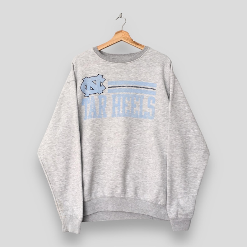 Op de afbeelding: Een grijze sweatshirt met een lichtblauw en zwart ontwerp. De sweatshirt heeft de tekst "TAR HEELS" in lichtblauw, met een logo boven de tekst. De sweatshirt heeft een ronde hals en lange mouwen.