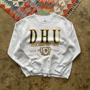 May include: A light gray sweatshirt with the text "DHU DUCK HEAD UNIVERSITY" in gold and green lettering. The sweatshirt also features a crest with a duck and the words "ESTABLISHED 1965".