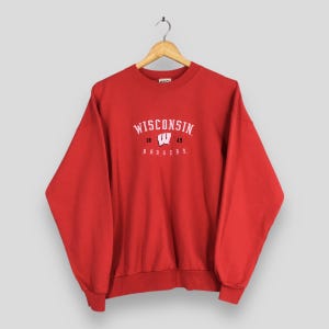 May include: A red sweatshirt with the word "WISCONSIN" arched across the chest in white lettering. Below the word "WISCONSIN" is the year "18 49" and the word "BADGERS" in white. The sweatshirt is hanging on a wooden hanger.