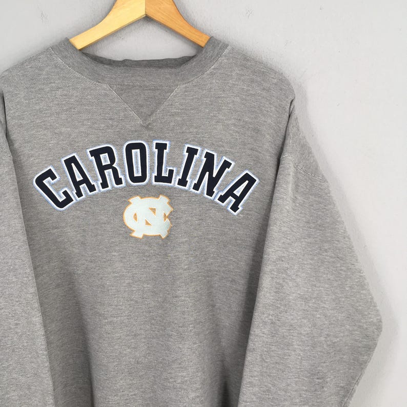 May include: A gray sweatshirt with the word "CAROLINA" in white and navy blue lettering. Below the word is a light blue and gold emblem. The sweatshirt is hanging on a wooden hanger.