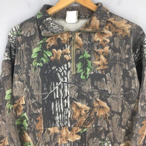 K&ouml;nnte beinhalten: Ein Sweatshirt mit Camouflage-Muster und einem Viertel-Rei&szlig;verschluss. Das Design enth&auml;lt braune, gr&uuml;ne und graue Elemente, die einer Waldszene &auml;hneln. Das Sweatshirt hat einen Kragen und lange &Auml;rmel.