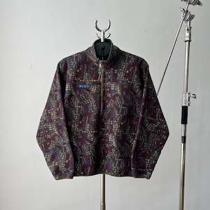 May include: A burgundy fleece pullover with a geometric pattern in beige, blue, and green. The long-sleeved top has a quarter-zip closure and a small logo patch. The fleece is hanging on a metal hanger.