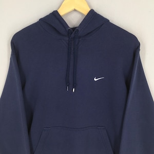 Pu&ograve; includere: Felpa con cappuccio blu navy con logo Nike bianco sul petto. La felpa ha un cappuccio con coulisse e una tasca a marsupio frontale. L'indumento &egrave; appeso a una gruccia di legno.