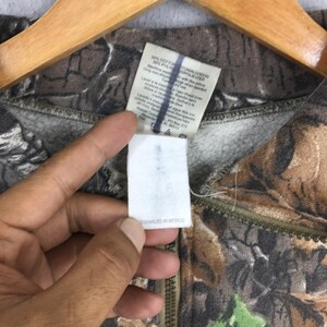 K&ouml;nnte beinhalten: Ein Sweatshirt mit Rei&szlig;verschluss und Camouflage-Muster. Der Stoff weist eine Mischung aus Braun-, Gr&uuml;n- und Beiget&ouml;nen auf. Ein wei&szlig;es Etikett mit Text ist sichtbar, zusammen mit einem Rei&szlig;verschluss. Das Etikett lautet "ASSEMBLED IN MEXICO".