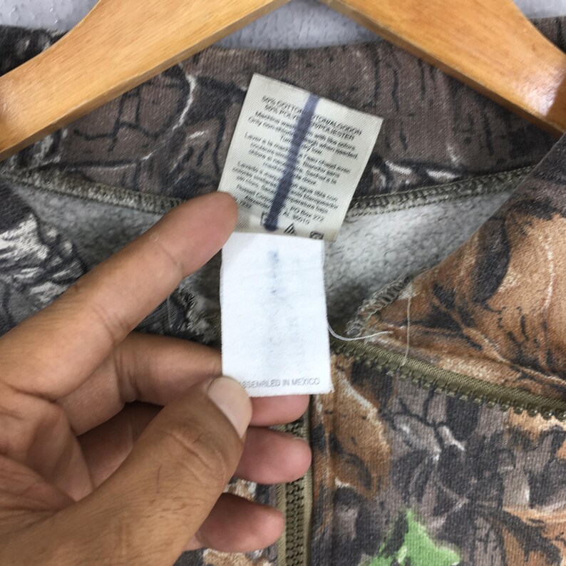 K&ouml;nnte beinhalten: Ein Sweatshirt mit Rei&szlig;verschluss und Camouflage-Muster. Der Stoff weist eine Mischung aus Braun-, Gr&uuml;n- und Beiget&ouml;nen auf. Ein wei&szlig;es Etikett mit Text ist sichtbar, zusammen mit einem Rei&szlig;verschluss. Das Etikett lautet "ASSEMBLED IN MEXICO".