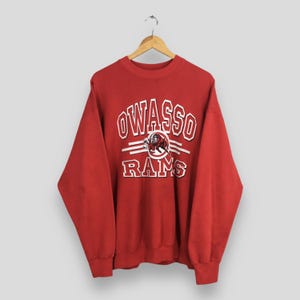 May include: A red sweatshirt featuring the word "OWASSO" in white block letters, with "RAMS" below. A ram graphic is in the center. The sweatshirt is suspended from a wooden hanger.