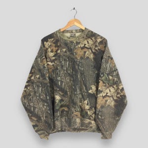Puede incluir: Sudadera de camuflaje de manga larga, con una mezcla de tonos marrones, verdes y beige. El diseño incorpora detalles de hojas y ramas, creando una estética de bosque. La sudadera está colgada de una percha de madera.