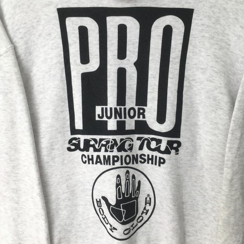 Puede incluir: Sudadera gris claro con un gran gr&aacute;fico negro que dice "PRO JUNIOR SURFING TOUR CHAMPIONSHIP". Debajo del texto hay un logotipo circular con una mano y las palabras "BODY GLOVE".