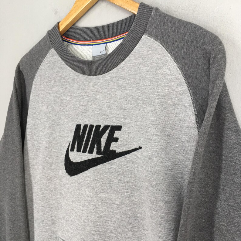 K&ouml;nnte beinhalten: Ein graues Nike-Sweatshirt mit Raglan&auml;rmeln. Das Sweatshirt hat ein gro&szlig;es schwarzes Nike-Logo auf der Vorderseite. Der Kragen hat ein gestreiftes Design mit dem Nike-Logo auf dem Kragen.