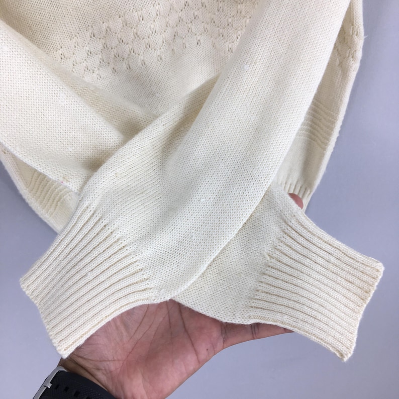 May include: Cream-colored knit sweater with ribbed cuffs and a diamond pattern on the upper chest. The sleeves are folded, showcasing the texture and detail of the fabric. The sweater appears soft and comfortable.