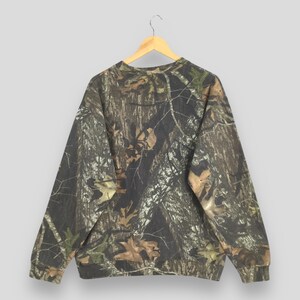 Puede incluir: Una sudadera con estampado de camuflaje cuelga de una percha de madera. El dise&ntilde;o presenta una mezcla de tonos marrones, verdes y beige, que imitan un entorno forestal. La prenda de manga larga tiene cuello redondo y pu&ntilde;os y dobladillo de canal&eacute;.