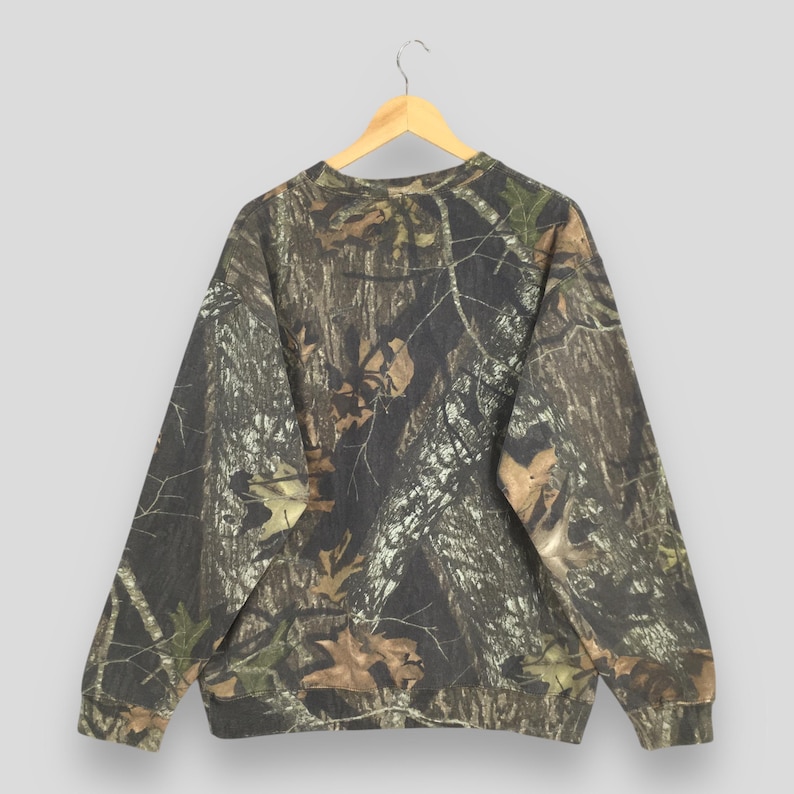 Puede incluir: Una sudadera con estampado de camuflaje cuelga de una percha de madera. El dise&ntilde;o presenta una mezcla de tonos marrones, verdes y beige, que imitan un entorno forestal. La prenda de manga larga tiene cuello redondo y pu&ntilde;os y dobladillo de canal&eacute;.