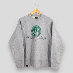 May include: A heather gray sweatshirt with a crew neck and long sleeves. The front features a green and white graphic of a hand inside a circle, with the words "Body Glove" in cursive below. The sweatshirt is hanging on a wooden hanger.
