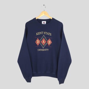 May include: Navy blue sweatshirt with the text "KENT STATE DAD UNIVERSITY" in gold, red, and green. The design features diamond shapes. The sweatshirt is hanging on a wooden hanger.