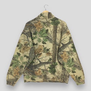 Puede incluir: Una sudadera con estampado de camuflaje y cuello alto, colgada de una percha de madera. El dise&ntilde;o presenta una mezcla de colores marr&oacute;n, verde y beige, que recuerda a una escena de bosque con hojas y ramas de &aacute;rboles. Se muestra la vista trasera de la sudadera.