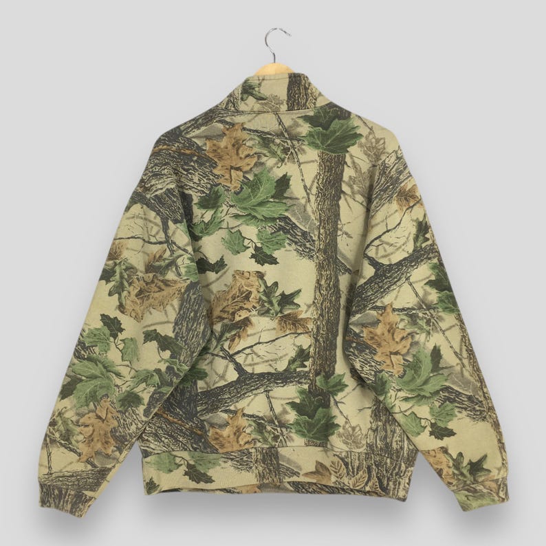 Puede incluir: Una sudadera con estampado de camuflaje y cuello alto, colgada de una percha de madera. El dise&ntilde;o presenta una mezcla de colores marr&oacute;n, verde y beige, que recuerda a una escena de bosque con hojas y ramas de &aacute;rboles. Se muestra la vista trasera de la sudadera.
