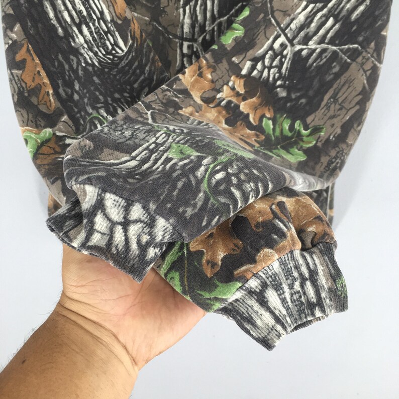 May include: Close-up of a camouflage-patterned sweatshirt sleeve. The fabric features a mix of brown, green, and gray tones, mimicking a forest environment. The cuff is ribbed and dark gray.