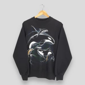 May include: Black sweatshirt with a detailed illustration of orcas and dolphins. The design features a large orca with a dolphin leaping above it, and two more orcas below. The artwork is in white, black, and blue, with water splashes.