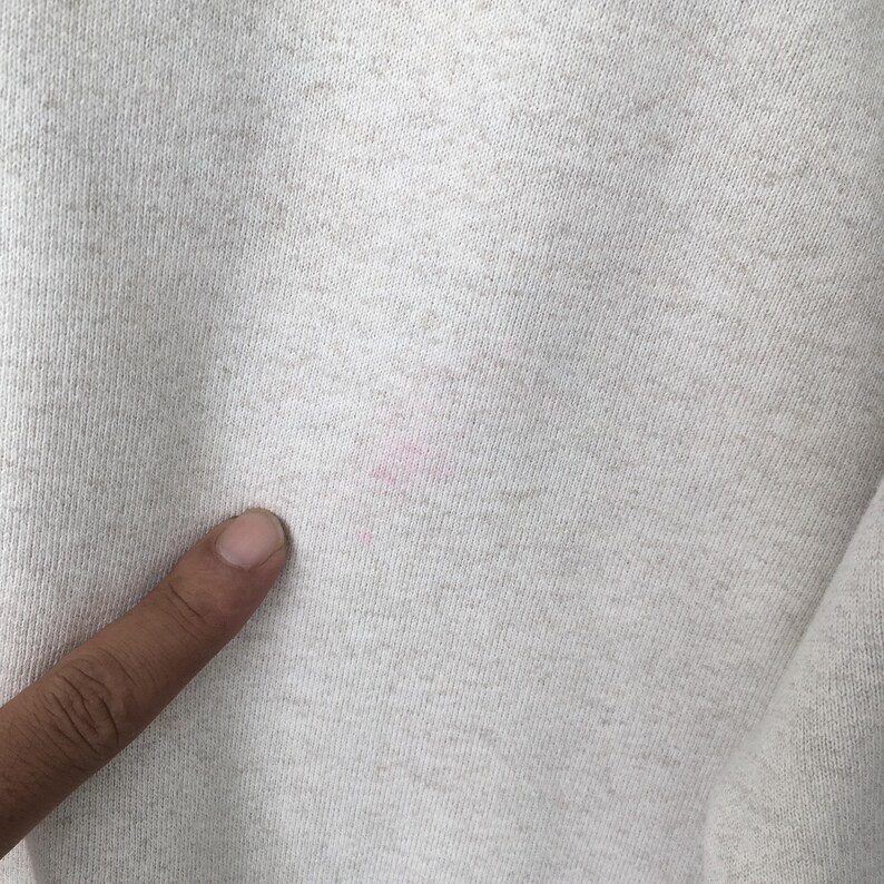 May include: Close-up of a light gray fabric with a textured surface. A pink stain is visible on the fabric. A finger is touching the fabric.