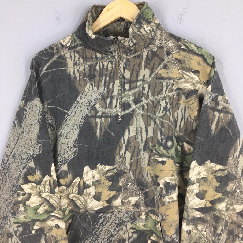 May include: A camouflage-patterned pullover sweatshirt with a half-zip closure. The design features shades of brown, green, and gray, mimicking a forest environment. The sweatshirt has a high collar and a front pocket.