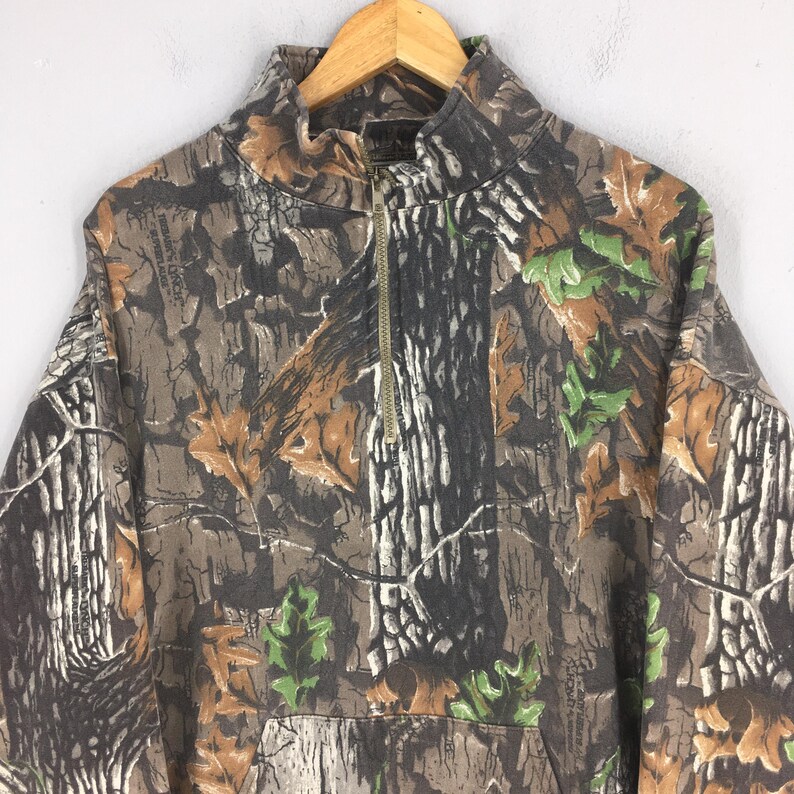 May include: A camouflage-patterned pullover sweatshirt with a high collar and a quarter-length zipper. The design incorporates brown, green, and gray tones, mimicking a forest environment. The sweatshirt is hanging on a wooden hanger.
