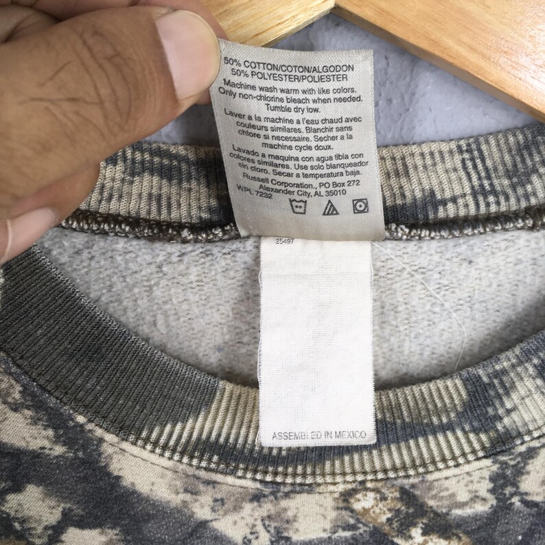 Peut inclure: Gros plan sur une &eacute;tiquette de v&ecirc;tement attach&eacute;e &agrave; un sweat-shirt &agrave; motif camouflage. L'&eacute;tiquette fournit des instructions de lavage en anglais, fran&ccedil;ais et espagnol, et des d&eacute;tails sur la composition du tissu. Le sweat-shirt est assembl&eacute; au Mexique.