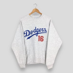May include: A grey marl sweatshirt featuring the word "Dodgers" in blue script and the number "16" in red. The sweatshirt is suspended on a wooden hanger against a plain backdrop.