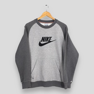 K&ouml;nnte beinhalten: Ein graues Nike-Sweatshirt mit dunkelgrauen Raglan&auml;rmeln und einer K&auml;ngurutasche. Das Nike-Logo ist schwarz auf der Brust. Das Sweatshirt h&auml;ngt an einem Holzb&uuml;gel.