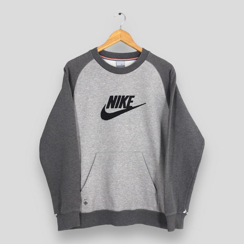K&ouml;nnte beinhalten: Ein graues Nike-Sweatshirt mit dunkelgrauen Raglan&auml;rmeln und einer K&auml;ngurutasche. Das Nike-Logo ist schwarz auf der Brust. Das Sweatshirt h&auml;ngt an einem Holzb&uuml;gel.