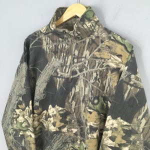 May include: Camouflage-patterned pullover sweatshirt with a quarter-zip closure. The fabric features a mix of brown, green, and tan hues, creating a woodland camouflage design. The sweatshirt has a high collar and long sleeves.