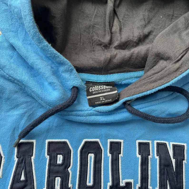 K&ouml;nnte beinhalten: Ein hellblauer Kapuzenpullover mit grauem Kapuzenfutter und Kordelzug. Das Wort "CAROLINA" ist in Marineblau auf die Vorderseite gestickt. Das Etikett lautet "Colosseum OWN THE STANDS" und "XL MADE IN CHINA".
