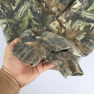 K&ouml;nnte beinhalten: Ein lang&auml;rmeliges Shirt mit Camouflage-Muster. Der Stoff zeigt eine Mischung aus Gr&uuml;n-, Braun- und Beiget&ouml;nen, die eine Waldumgebung nachahmen. Das &Auml;rmelb&uuml;ndchen ist sichtbar und zeigt die gerippte Textur und Farbvariationen des Camouflage-Designs.
