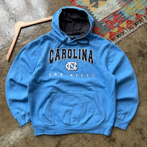 K&ouml;nnte beinhalten: Hellblaues Kapuzen-Sweatshirt mit dem Wort "CAROLINA" in Marineblau und Wei&szlig; &uuml;ber der Brust. Darunter befindet sich ein wei&szlig;es und marineblaues Logo. Die Worte "TAR HEELS" stehen darunter. Das Sweatshirt hat eine Vordertasche.