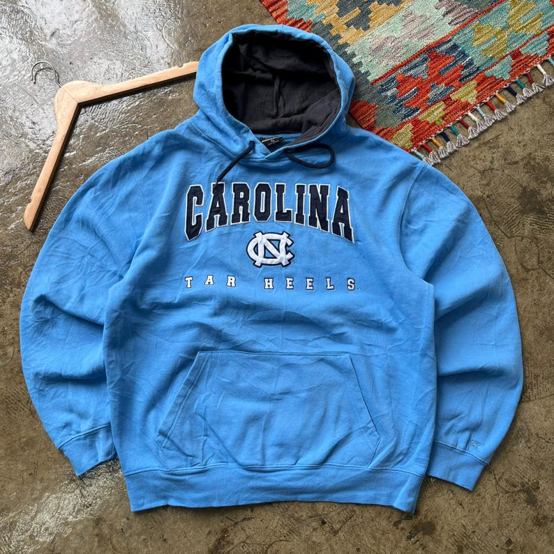 K&ouml;nnte beinhalten: Hellblaues Kapuzen-Sweatshirt mit dem Wort "CAROLINA" in Marineblau und Wei&szlig; &uuml;ber der Brust. Darunter befindet sich ein wei&szlig;es und marineblaues Logo. Die Worte "TAR HEELS" stehen darunter. Das Sweatshirt hat eine Vordertasche.