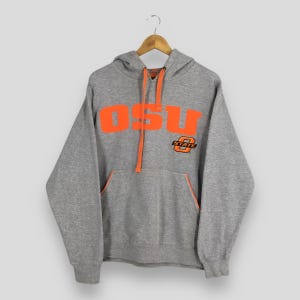 May include: A gray hoodie with the letters "OSU" in bright orange across the chest. The hoodie has a kangaroo pocket and orange drawstrings. A small "STATE" logo is on the right side. The hoodie is hanging on a wooden hanger.