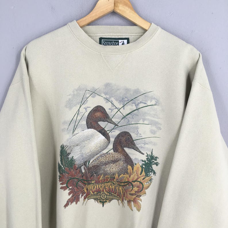 Peut inclure: Un sweat-shirt beige avec un imprim&eacute; de deux canards dans un marais. Le texte "The Sportsman" est imprim&eacute; sous les canards.