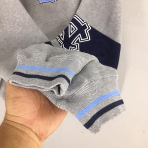 Puede incluir: Primer plano de una manga de sudadera gris con rayas azules y marino en el pu&ntilde;o. La manga tiene un panel azul marino con letras blancas. La sudadera es de estilo deportivo.
