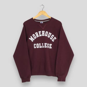 Puede incluir: Una sudadera de cuello redondo y manga larga en color granate con el texto blanco "MOREHOUSE COLLEGE" arqueado en el pecho. La prenda incluye cuello, puños y dobladillo acanalados, con un pequeño detalle de costura triangular bajo el cuello.