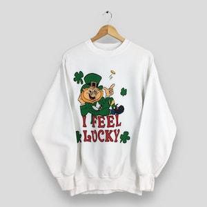 Op de afbeelding: Witte sweatshirt met een cartoon kabouter met een groene hoed en pak, omringd door klavers. De tekst "I FEEL LUCKY" is in rood en groen gedrukt. Een gouden munt zweeft boven de vinger van de kabouter.