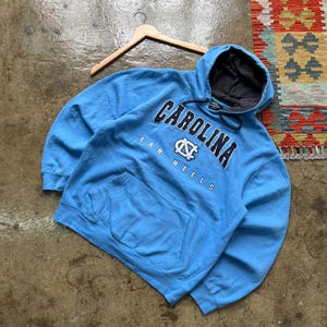 K&ouml;nnte beinhalten: Hellblauer Kapuzenpullover mit dem Wort "CAROLINA" in marineblauen Buchstaben auf der Brust. Unter dem Wort "CAROLINA" steht der Schriftzug "TAR HEELS" und ein Logo. Der Hoodie hat eine Vordertasche und ein graues Kapuzenfutter.