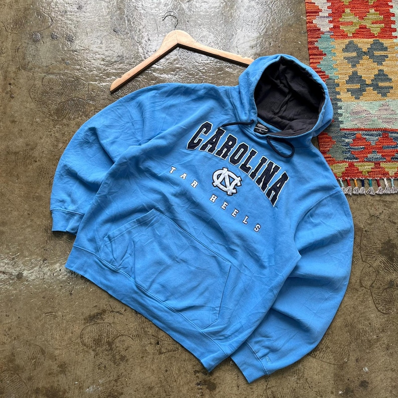 K&ouml;nnte beinhalten: Hellblauer Kapuzenpullover mit dem Wort "CAROLINA" in marineblauen Buchstaben auf der Brust. Unter dem Wort "CAROLINA" steht der Schriftzug "TAR HEELS" und ein Logo. Der Hoodie hat eine Vordertasche und ein graues Kapuzenfutter.