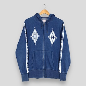 May include: A blue denim zip-up hoodie with a white geometric design on the front and sleeves. The sleeves have the text "RODEO CROWNS" printed vertically in white. The hoodie has a front pocket and a hood.