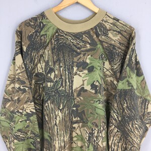 Op de afbeelding: Een sweatshirt met camouflagepatroon met een beige ronde hals en raglanmouwen. Het ontwerp heeft een mix van bruine, groene en beige tinten, die een bosrijke omgeving nabootsen. De sweatshirt hangt aan een houten hanger.