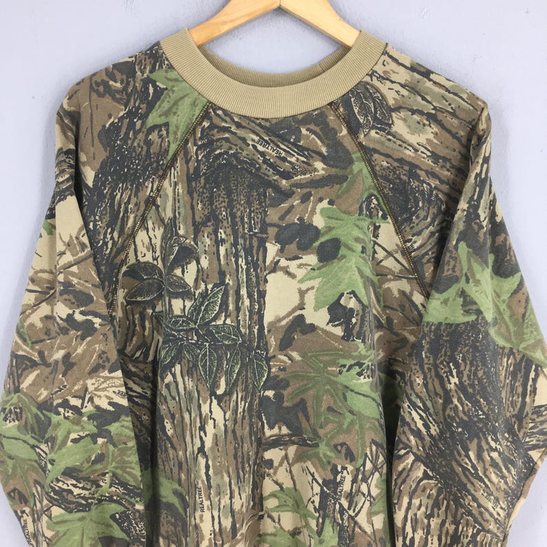 Op de afbeelding: Een sweatshirt met camouflagepatroon met een beige ronde hals en raglanmouwen. Het ontwerp heeft een mix van bruine, groene en beige tinten, die een bosrijke omgeving nabootsen. De sweatshirt hangt aan een houten hanger.